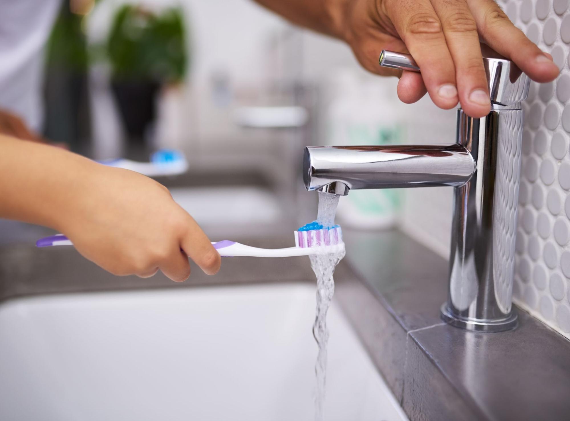 Close-up of a child rinsing a toothbrush under running water at the bathroom sink.