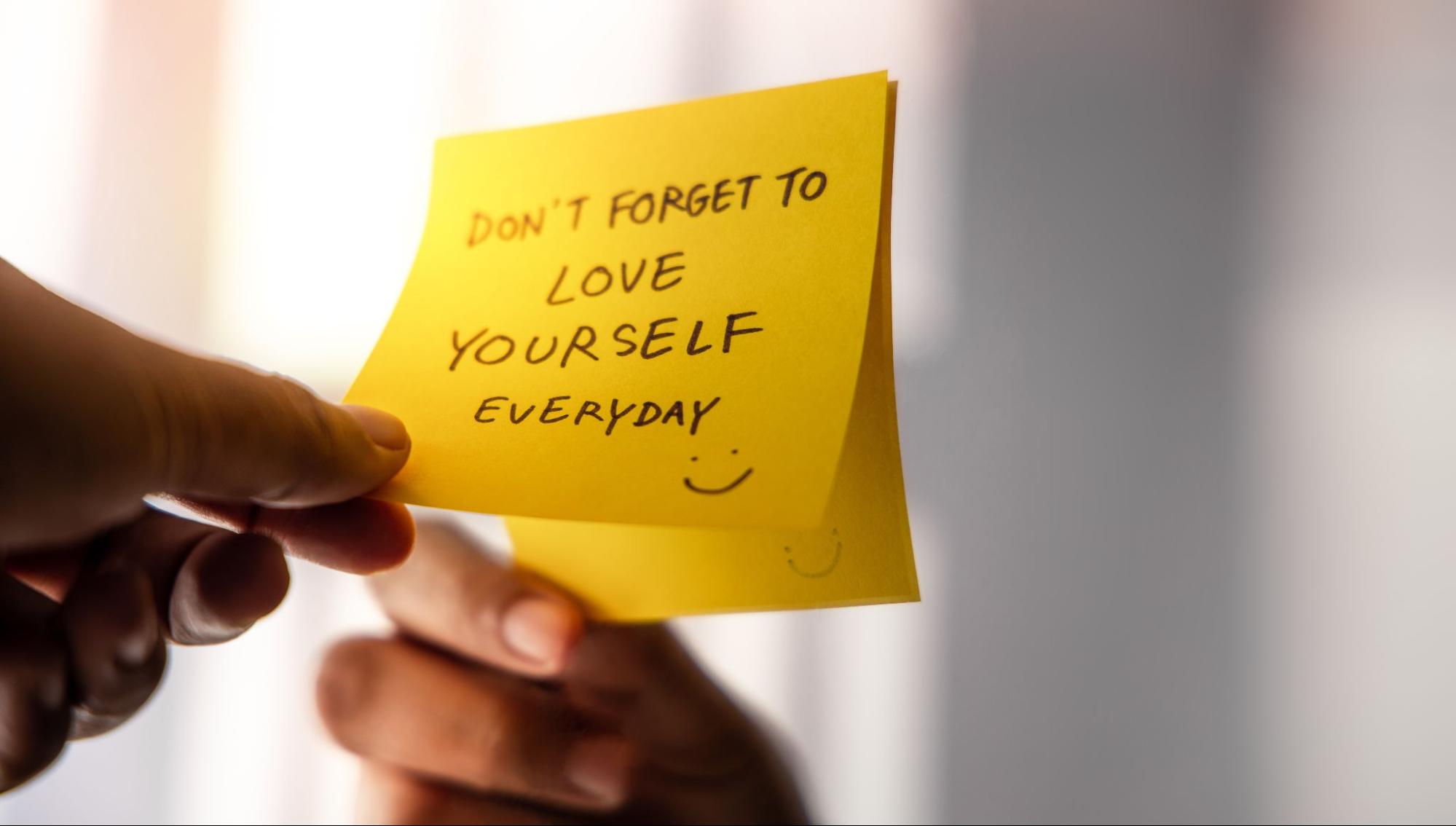 hand holding a sticky note with the message "Don't forget to love yourself everyday" for a mental health reminder.