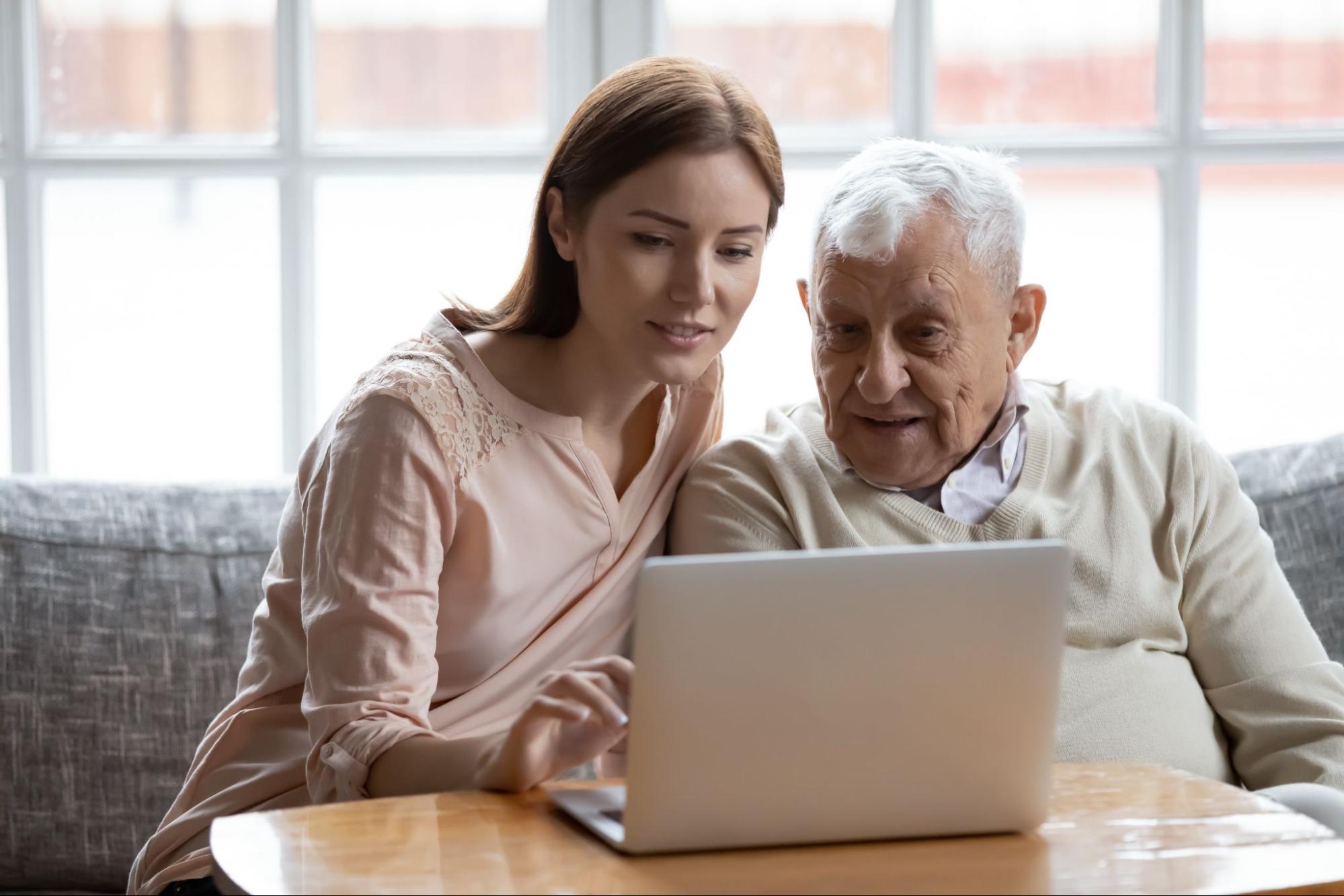 Young woman assisting an older man with a telehealth appointment on a laptop.