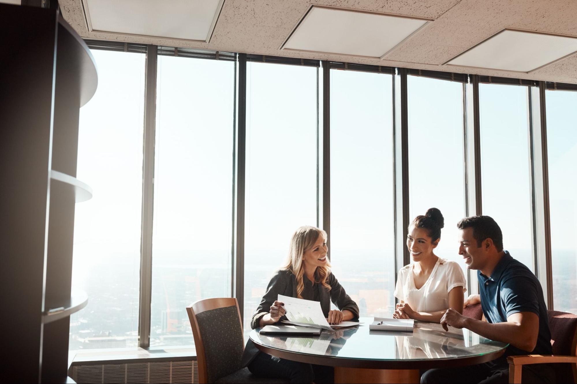 A professional advisor meets with a smiling couple in a bright office, discussing documents together at a round glass table.